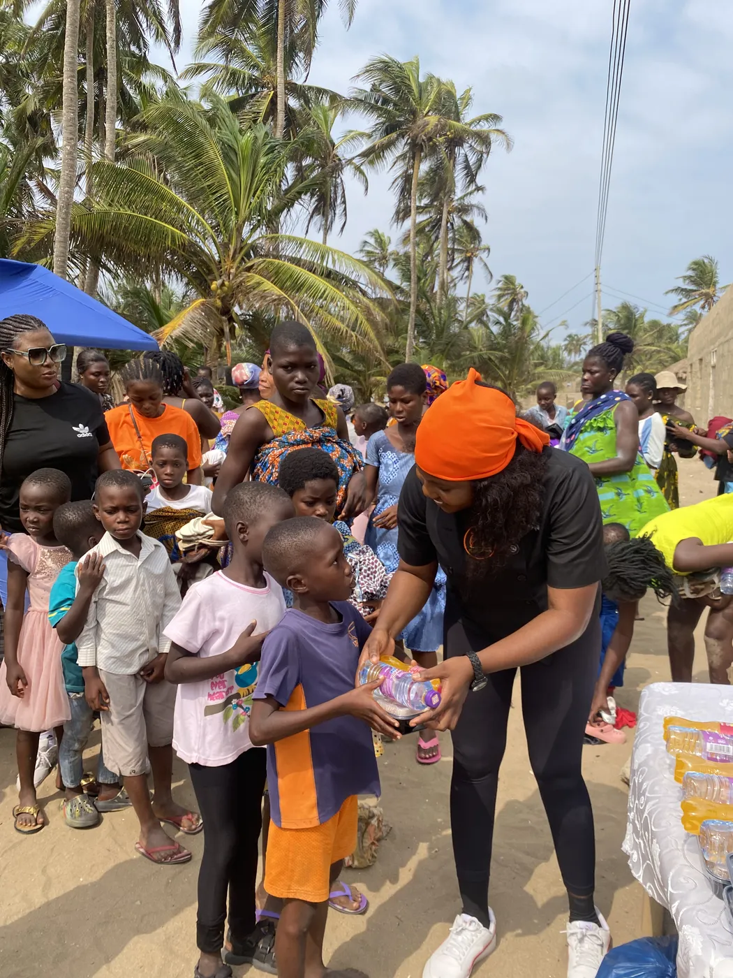 Chef Maamle in an orange headwrap handing an item to a child among a crowd of children under palm trees